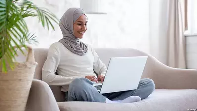 A smiling Muslim woman wearing a hijab sits and looks at her laptop