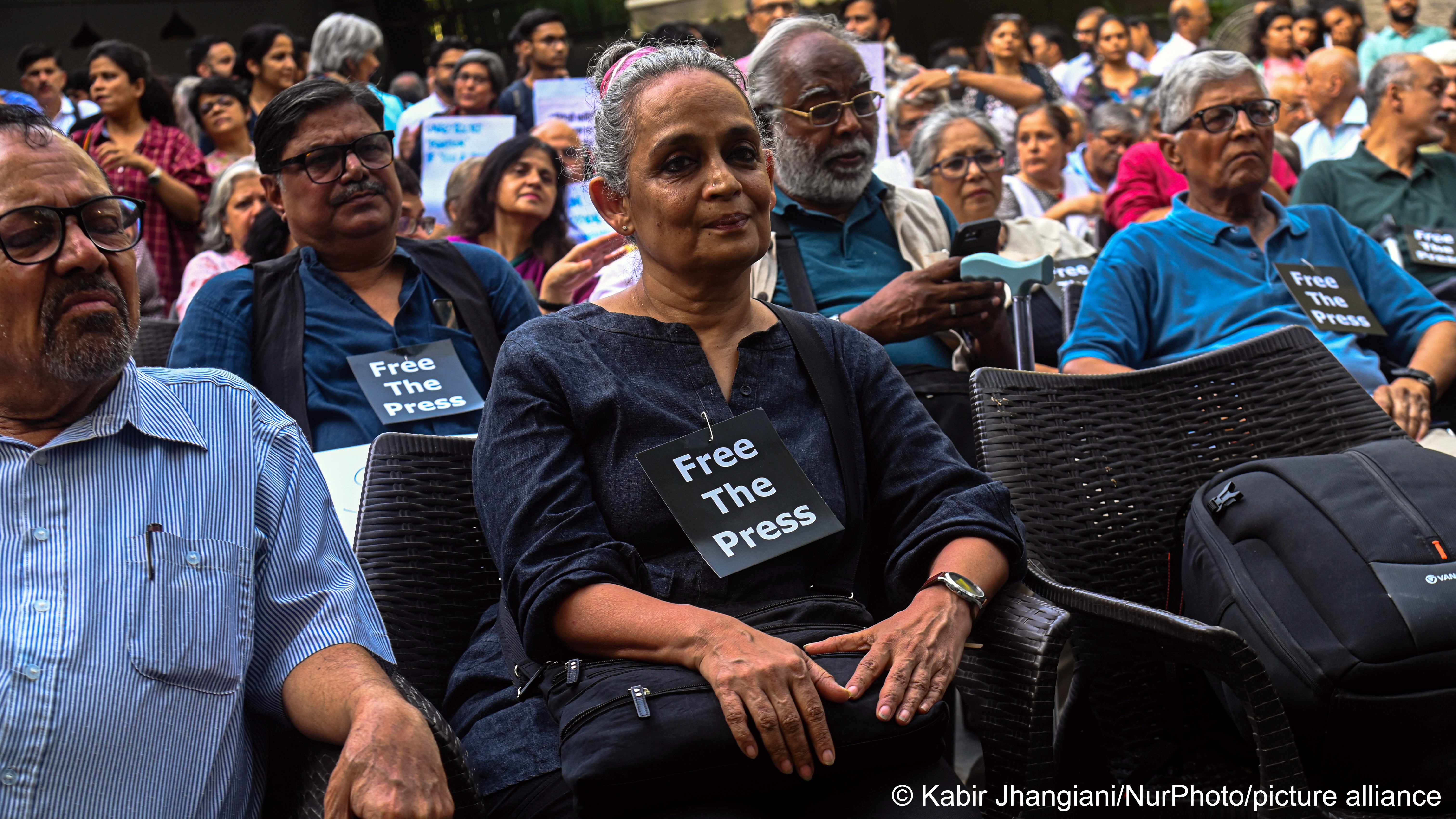 Prize-winning Indian author Arundhati Roy sits in a gathering of people with a sign round her neck bearing the words "Free the press"