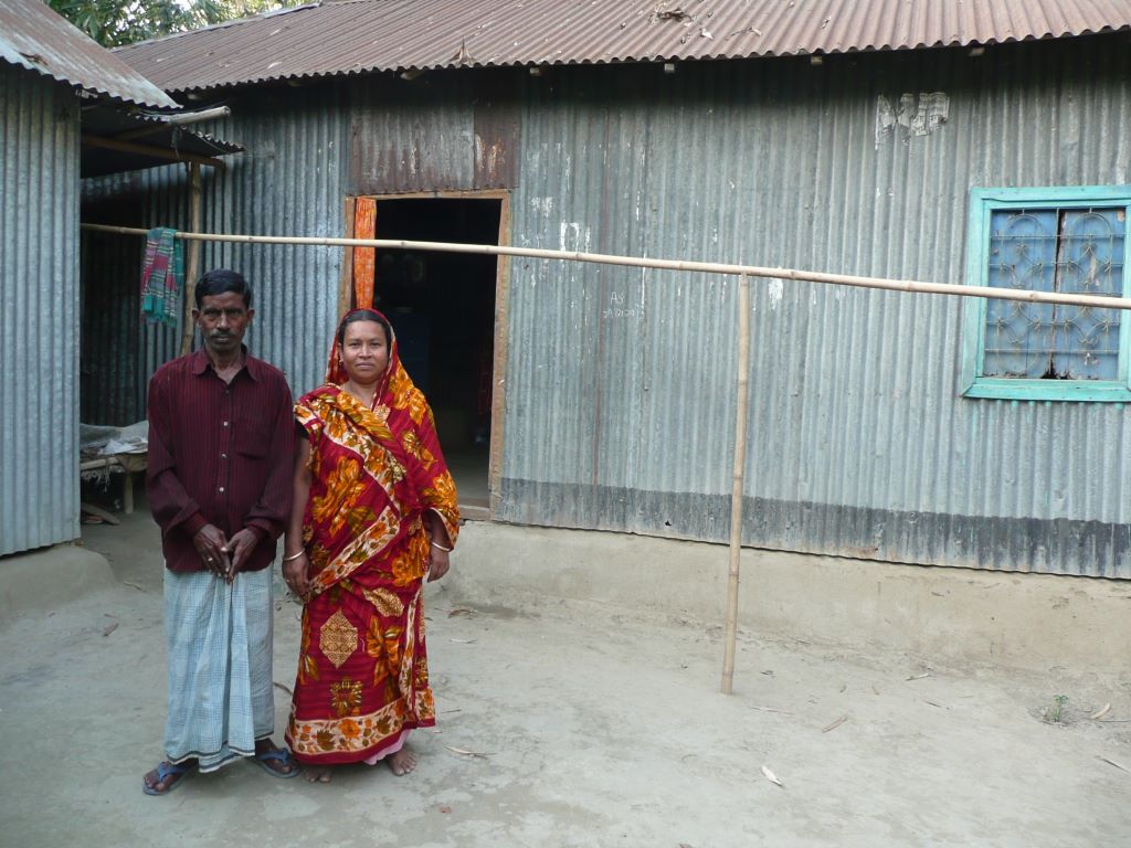 Rikha Begum and her husband stand outside a one-storey building in Bangladesh