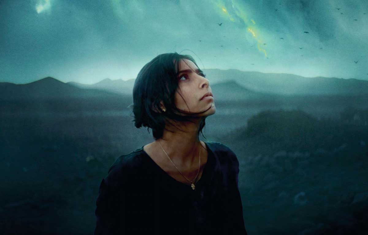 A woman in dark clothes looks up at a lowering sky against a background of hills obscured by haze