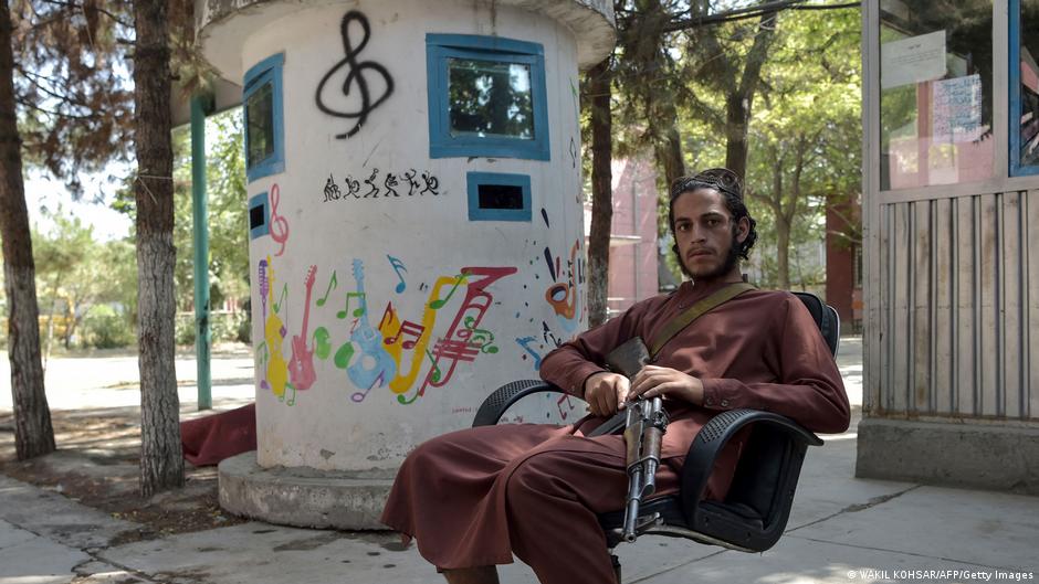 مسلح من حركة طالبان عند مدخل معهد الموسيقى الوطني في أفغانستان عام 2021.  ‏image: AFP/Getty Images An armed Taliban fighter lounges in a chair at the entrance to the Afghanistan National Institute of Music compound in September 2021; a treble clef graffito can be seen on the pillar behind him