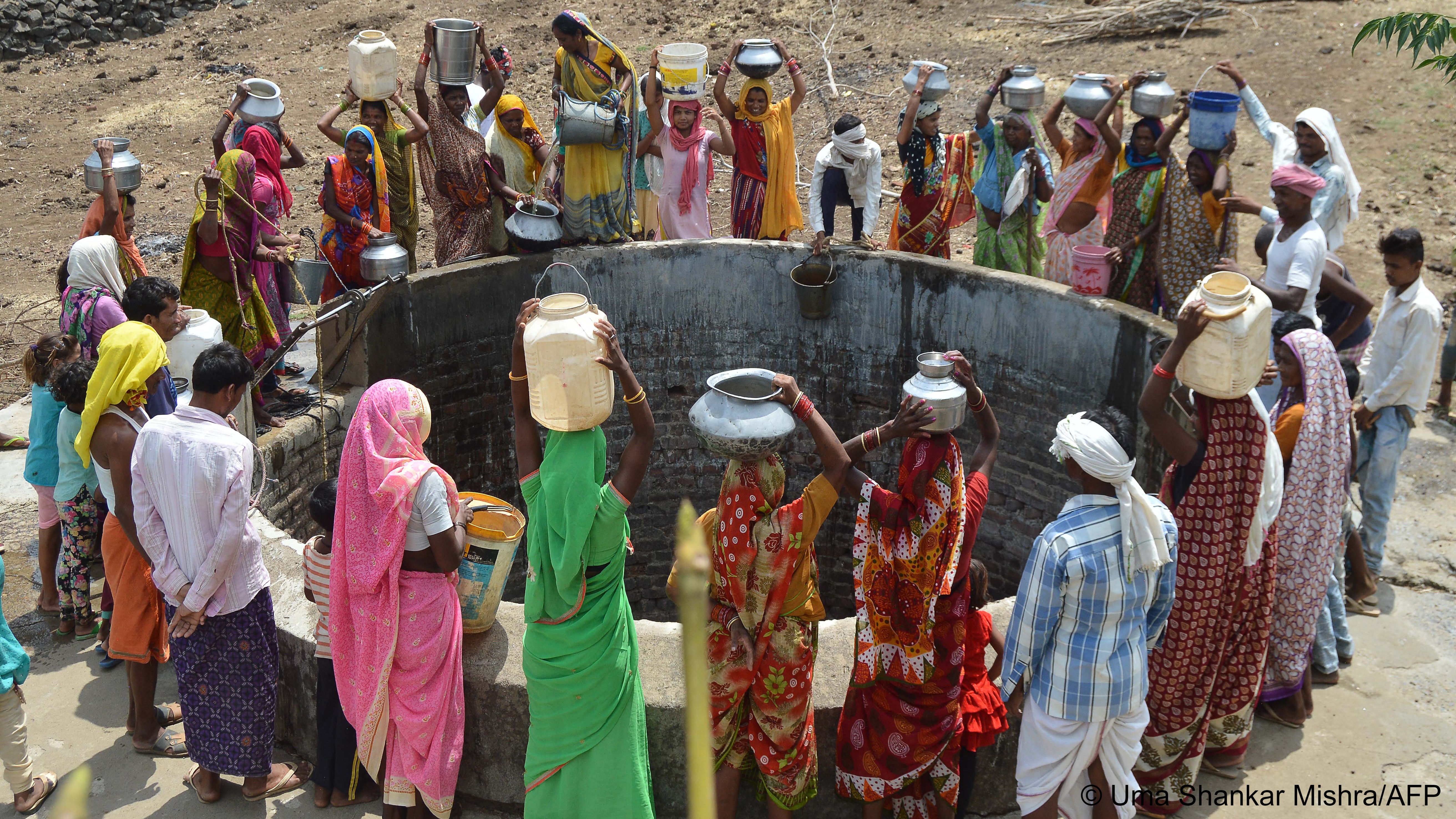 Women with water containers stand around a large round brick-built well