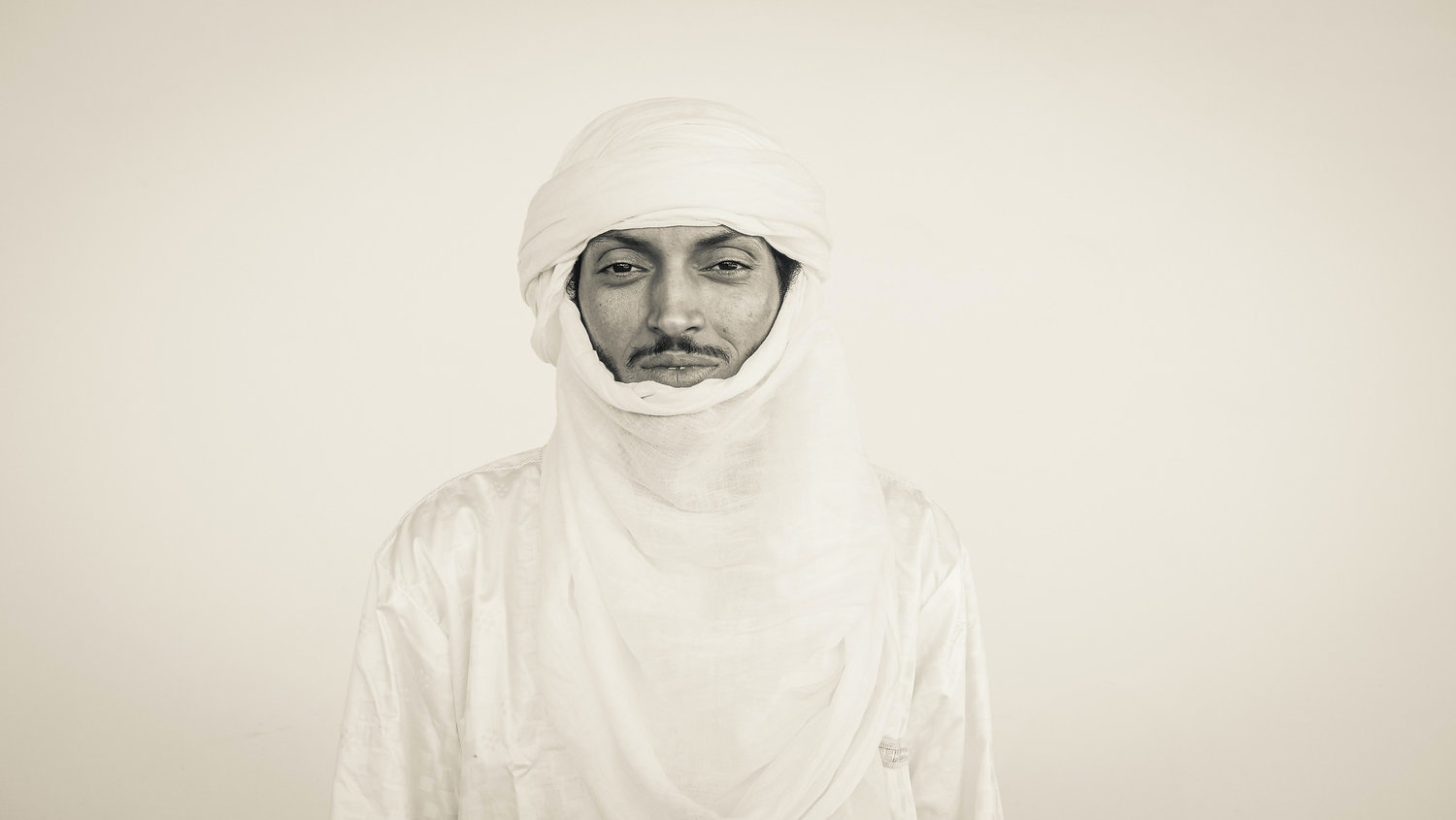 A man (Nigerien Musician Oumara Moctar/Bombino) dressed entirely in a white traditional Tuareg outfit and headdress against a white backdrop