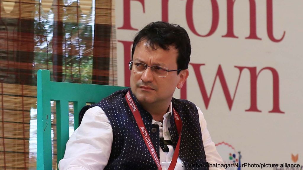 Indian art critic Ranjit Hoskote sits on a turquoise chair, a striped curtain and a white wall bearing large red letters behind him