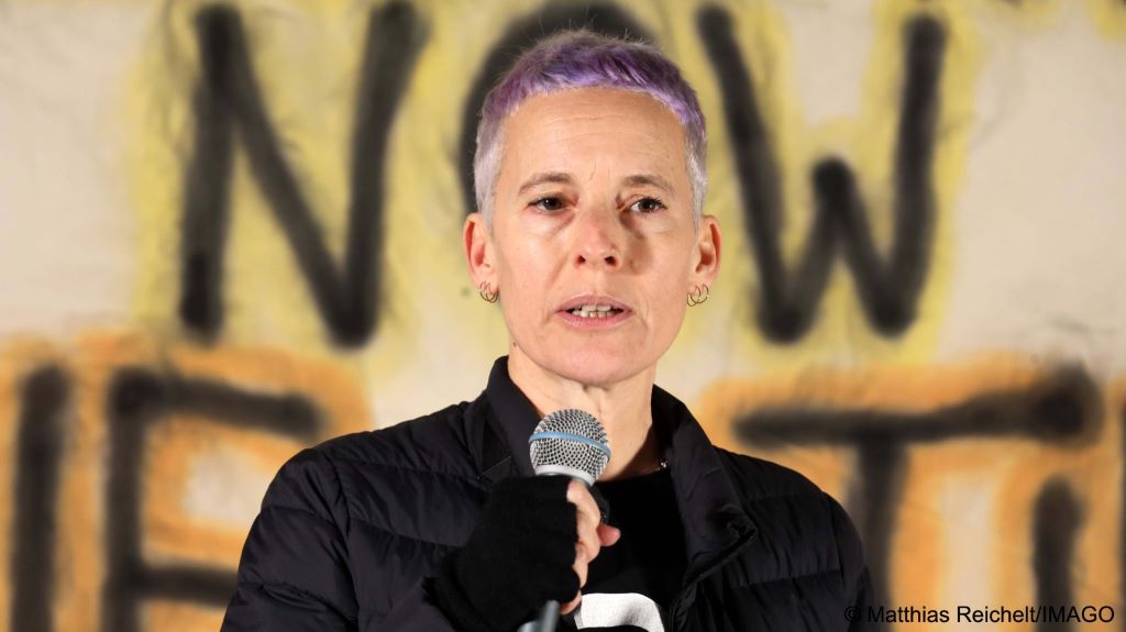 Jewish South African artist Candide Breitz, with short purple tinted hair, speaks into a microphone; large letters in black are seen in the background