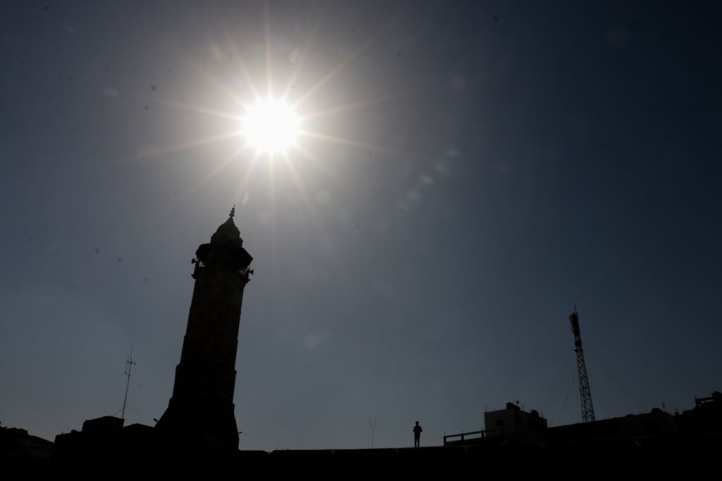The silhouette of a tower, roofs, pylon and a person can be seen against the sun