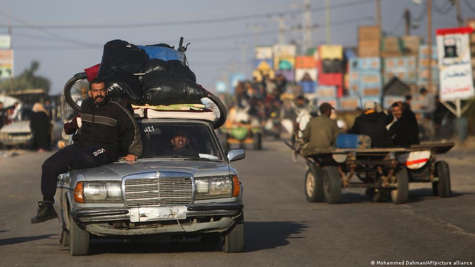 A overloaded car, its roof packed high with luggage and a man perched on its bonnet, drives along a street in Khan Younis. A donkey cart transports people in the other direction