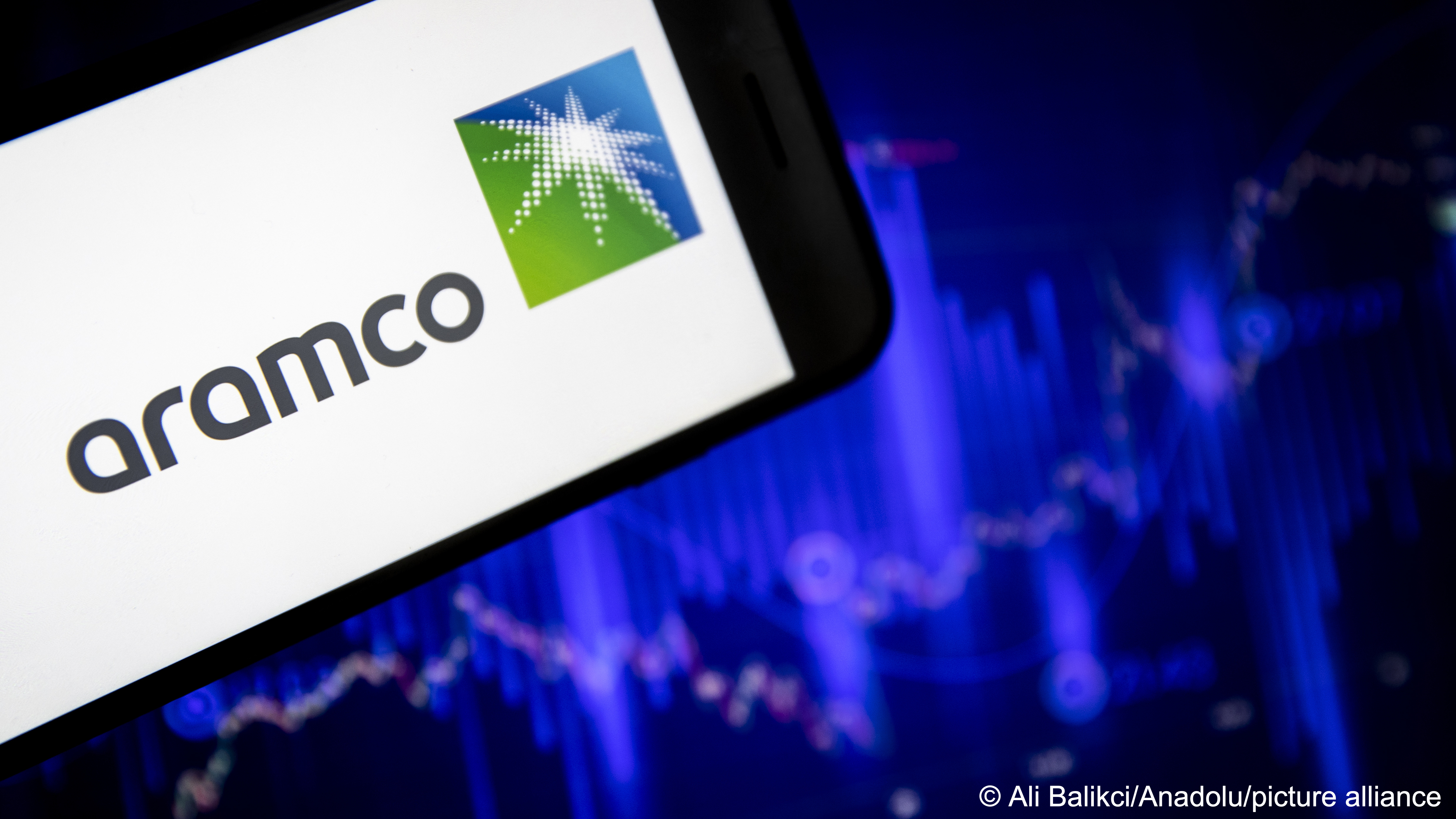 The logo of Saudi Aramco, the national oil and gas company of Saudi Arabia, is displayed on a mobile phone screen in front of a financial stock market graph