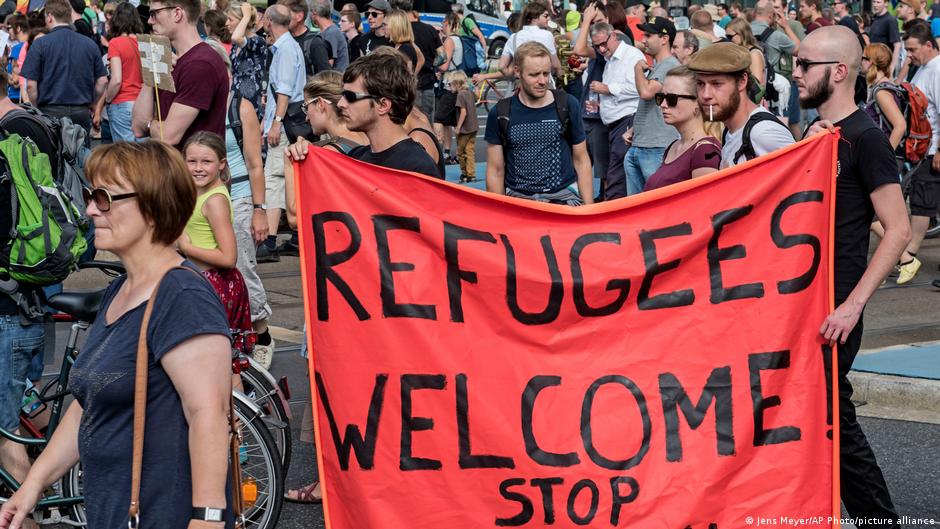 Two men carry a red banner reading "Refugees Welcome" through a crowd of people
