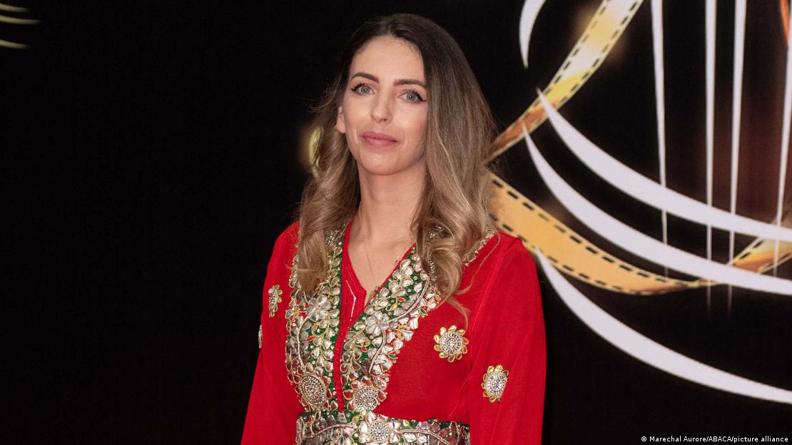 Film director Sofia Alaoui in a red silk dress with elaborate embroidery, against a black background with a gold and white design