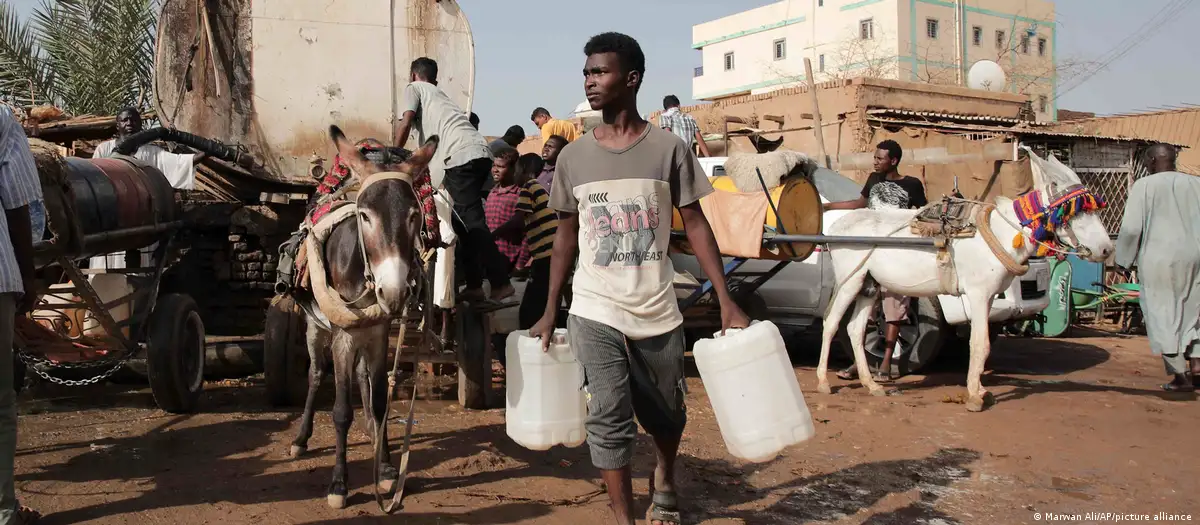 A man carrying two white canisters walks towards the camera, behind him are a crowd of people, carts and donkeys