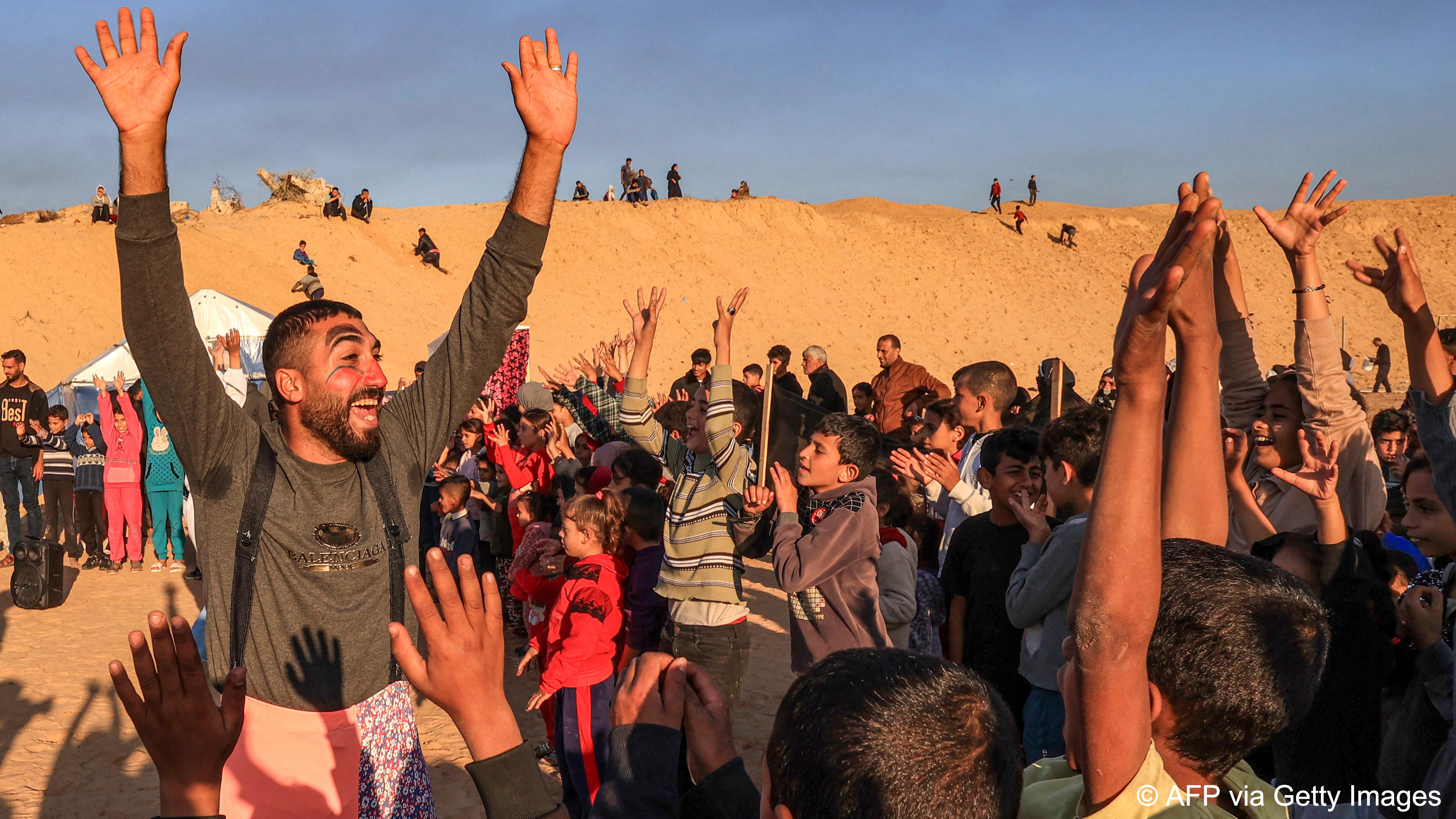 A man wearing clown make-up grins broadly as he raises his arms in the air and entertains a crowd in a camp near Rafah
