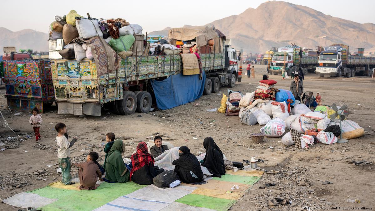 Afghan refugees sit on a mat on the ground. In the background are several trucks packed with the refugees' personal belongings 