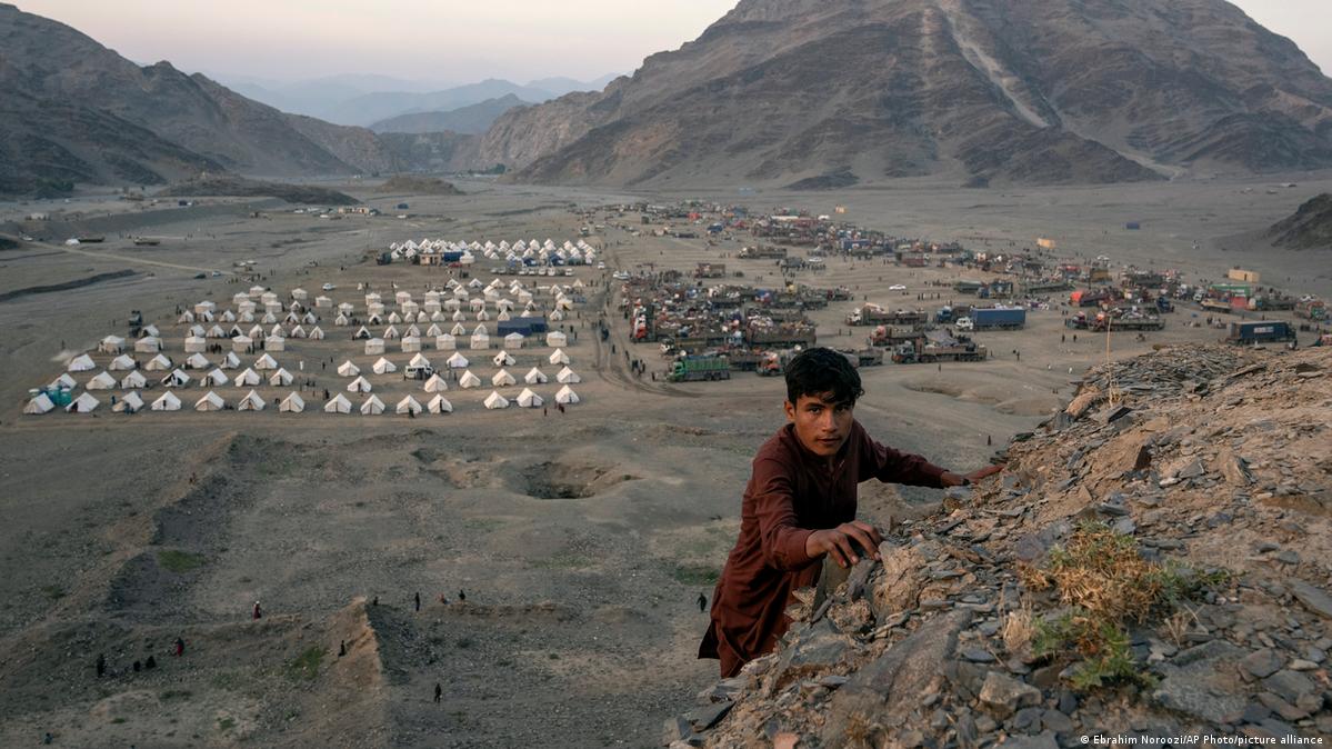 An Afghan boy climbs up a hill. In the background are rows of tent camps and fully packed, parked trucks in the valley below 