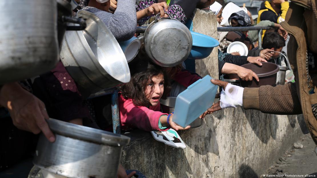 A crowd of people, including a young girl and a young boy, hold out empty pots and bowls through railings in the hope of getting food from a charity kitchen in Rafah, Gaza, February 2024