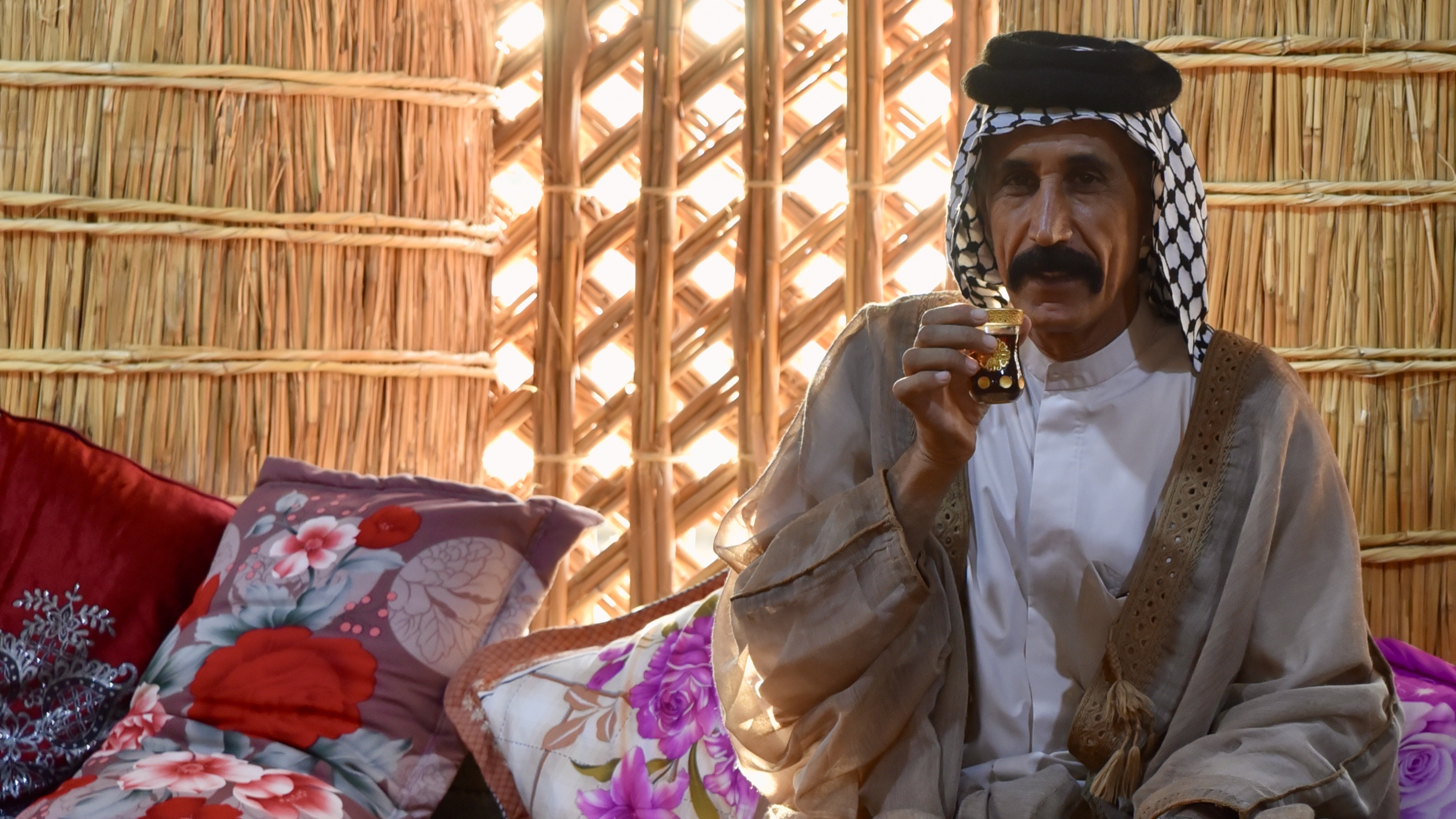 A Marsh Arab in traditional dress sips tea from a glass in a mudhif