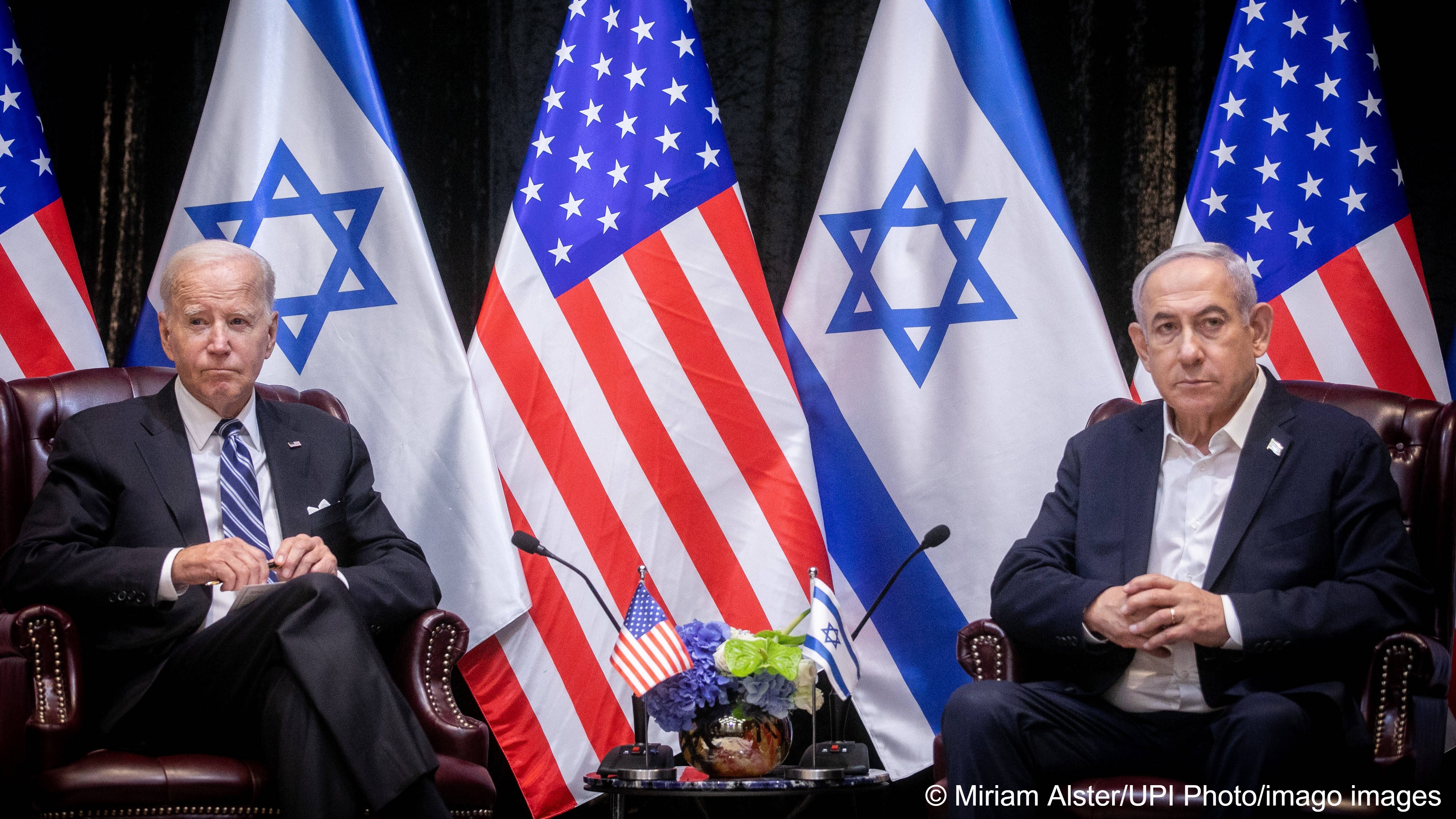 US President Joe Biden (left) and Israeli Prime Minister Benjamin Netanyahu (right) sit in armchairs in front of a row of American and Israeli flags, Tel Aviv, Israel, 18 October  2023
