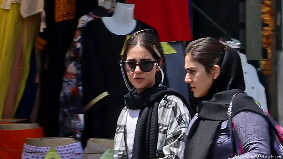 Young women walk along a street in Iran, one with her headscarf half covering her hair, one with her headscarf completely down