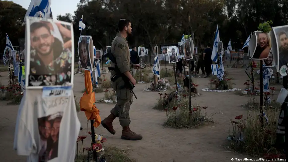 A soldier with a rifle over his shoulder walks through a makeshift memorial to the people massacred by Hamas at the Supernova music festival in Israel on 7 October. There are flowers and photos, Israeli flags and items of clothing on poles