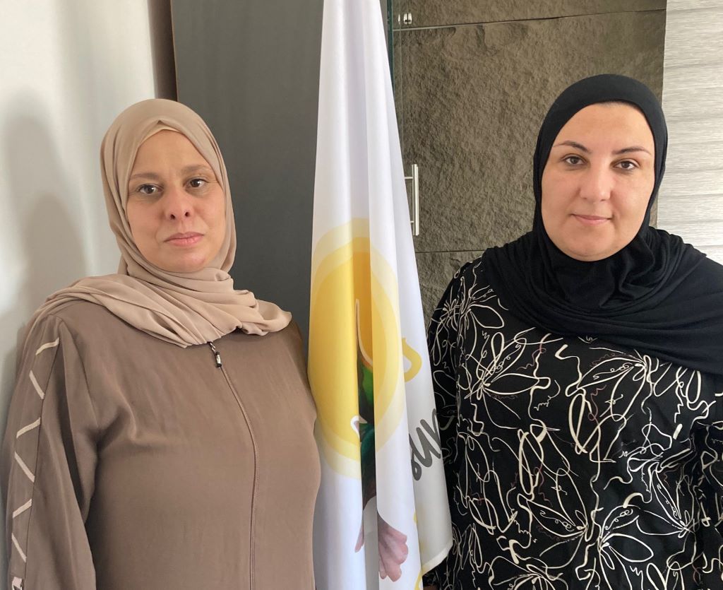 Two women in headscarves, Marwa Hamad (left) and Reem Alhajajreh stand on either side of a flag bearing the logo of the Women of the Sun NGO