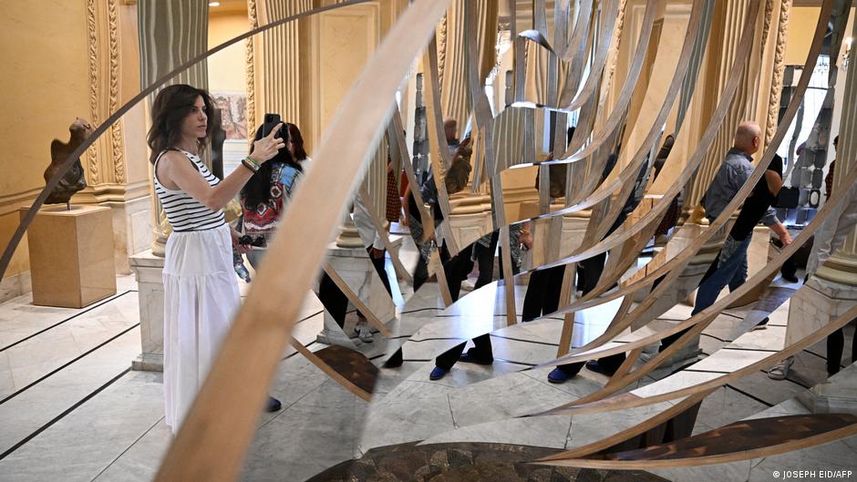 A woman uses her smartphone to take a photo of art as she stands behind a sweeping mirror installation with arched mirrored arms