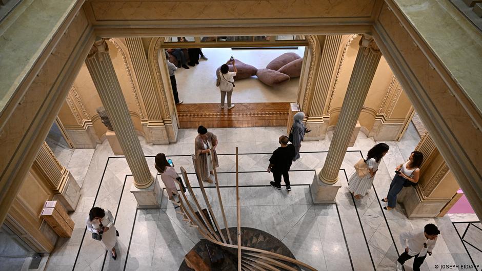 Visitors to an artistic expo seen from above as they stand and walk through a marble-floored and colannaded gallery with a large circular sculpture in the middle