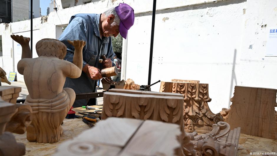 A man uses wood-working tools to work on wood creations while being surrounded by wood-carved sculptures