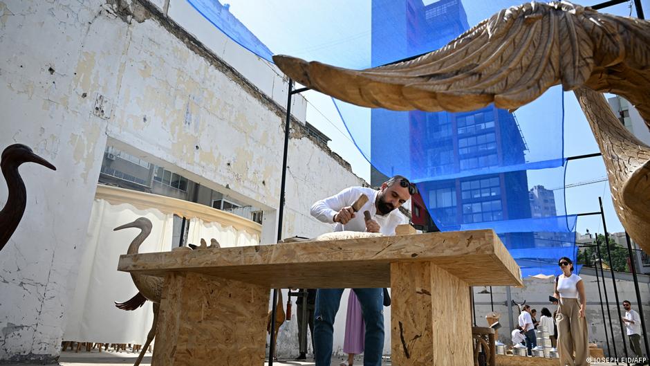 Against the backdrop of a tall building and a long low building, a man uses wood-working tools to work on something on top of a large wooden table