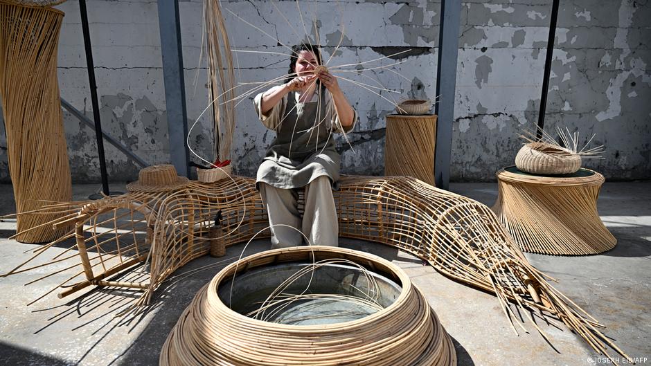 A woman holds rattan palm strands in the air and weaves them together while sitting on another larger curved rattan bench structure