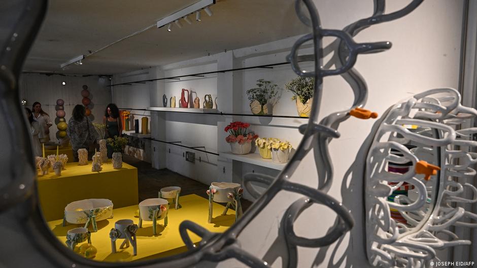 A mirror with a twisted wire-like frame reflects an exhibition room filled with small sculptures and vases on tables and wall shelves 
