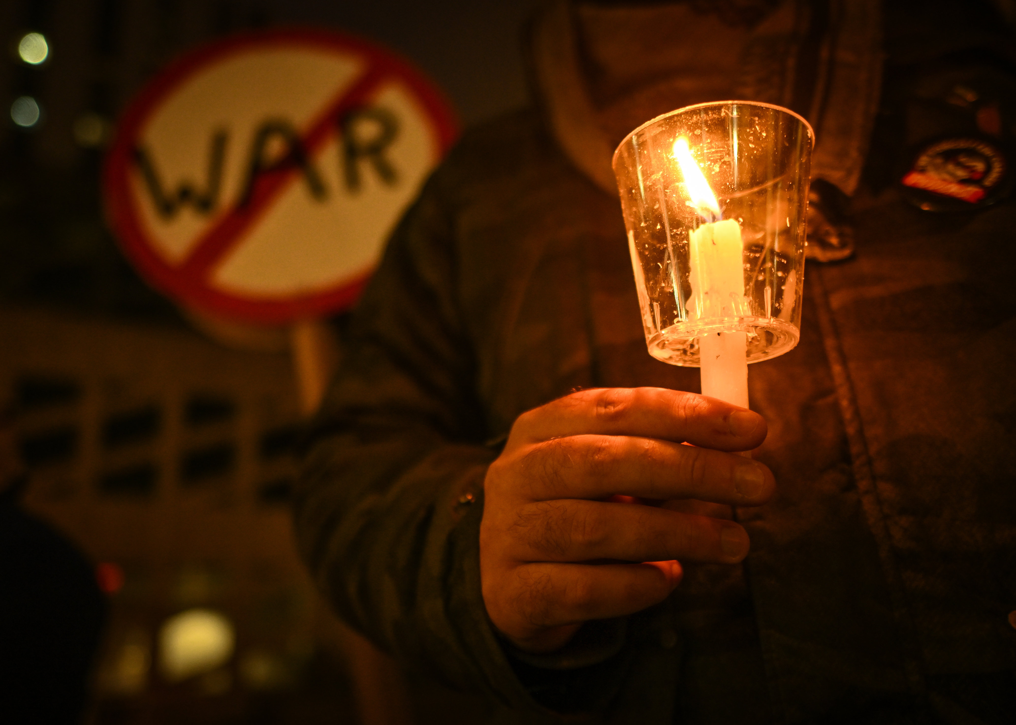 Nahaufnahme eines Menschen, der eine Kerze hält. Hinter ihm ein Schild auf dem durchgestrichen "Krieg" steht