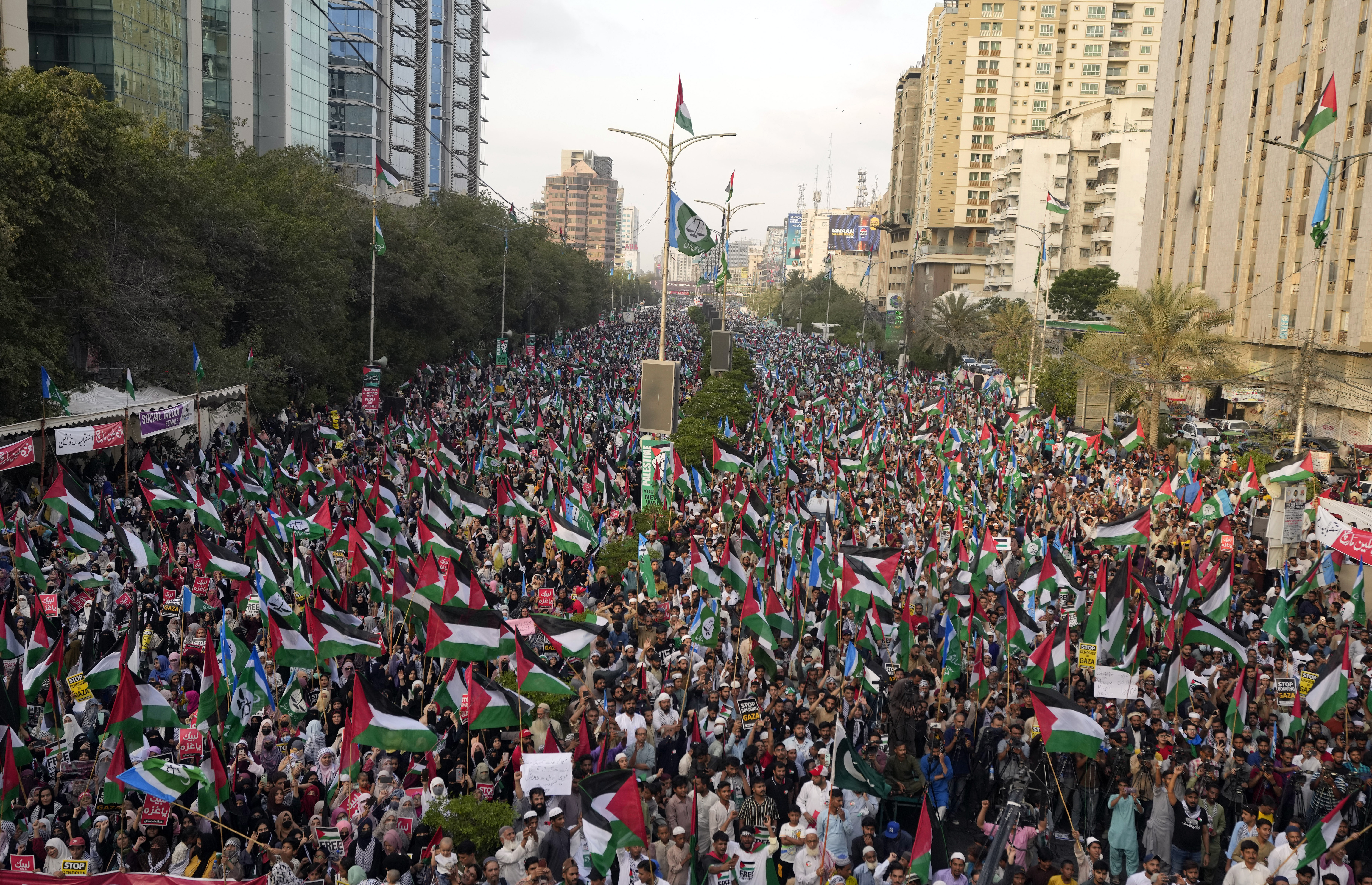 A large crowd of people holding Palestine flags and signs walk down a wide road in Karachi.