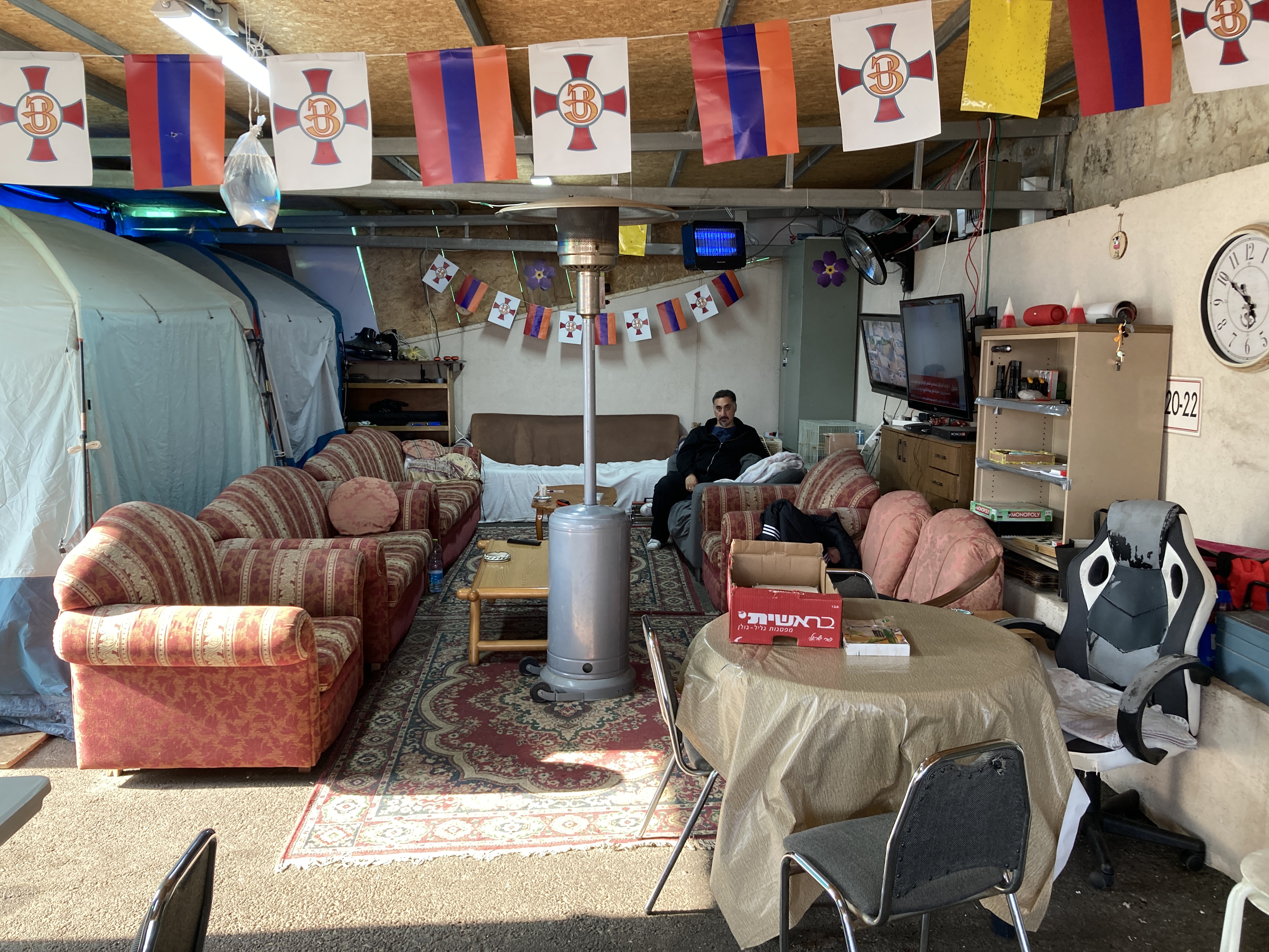 A man sits alone on a sofa inside a camp roughly constructed from pieces of board, tents and other materials.