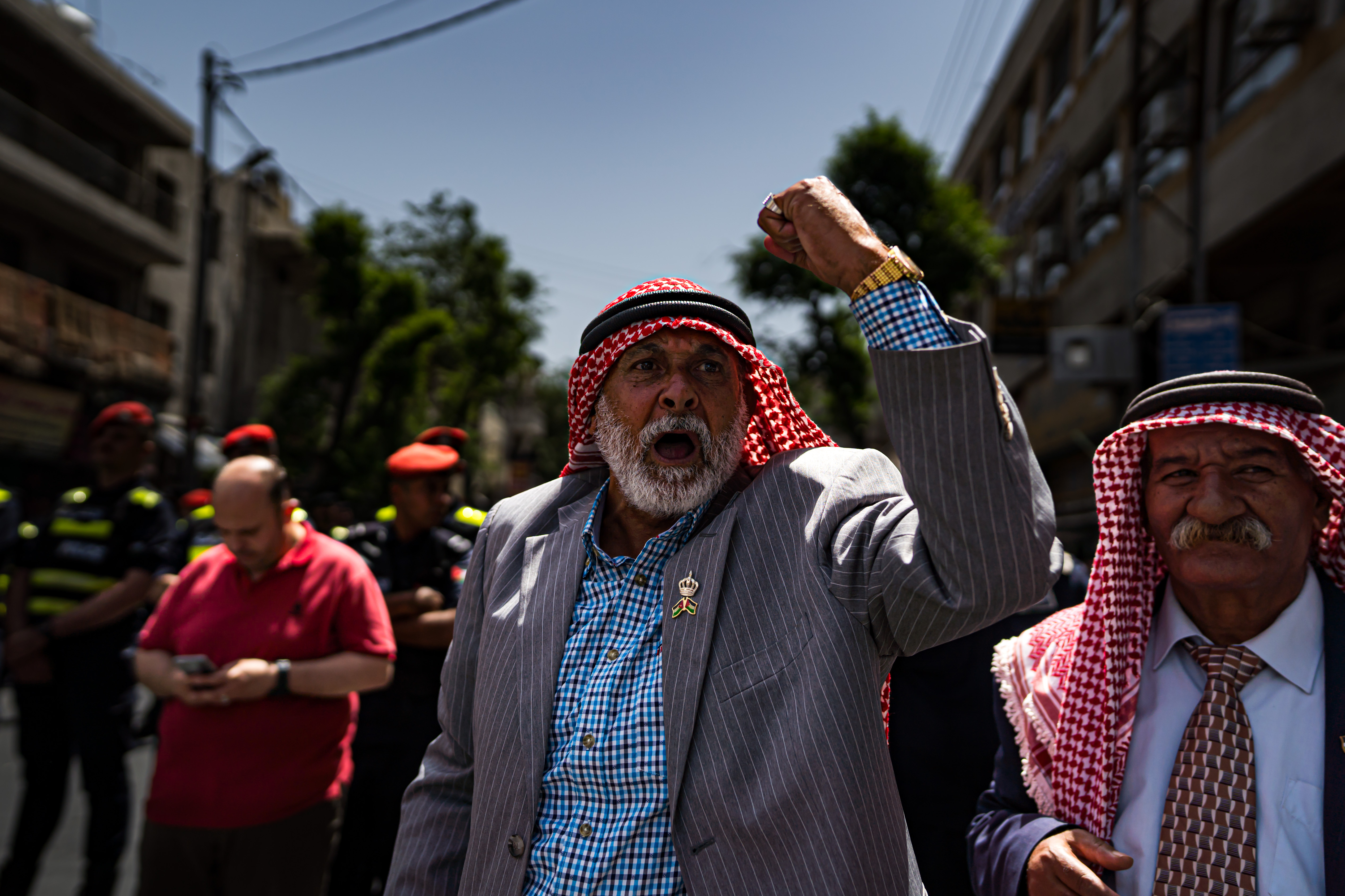 In the foreground are two men wearing suits and red keffiyehs. One of them raises his fist to the sky and shouts.