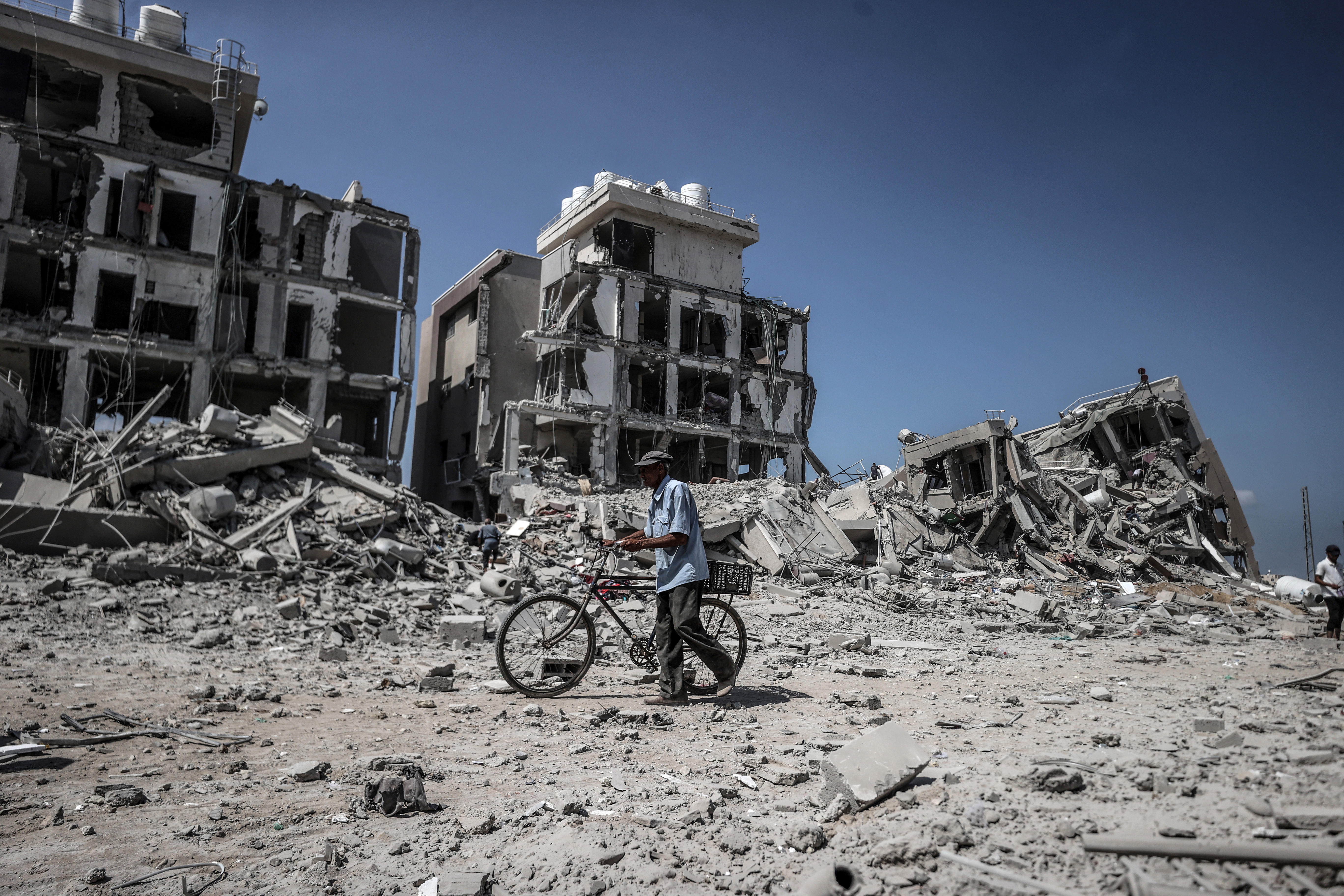 A man pushes a bicycle through rubble. In the background are three destroyed buildings.