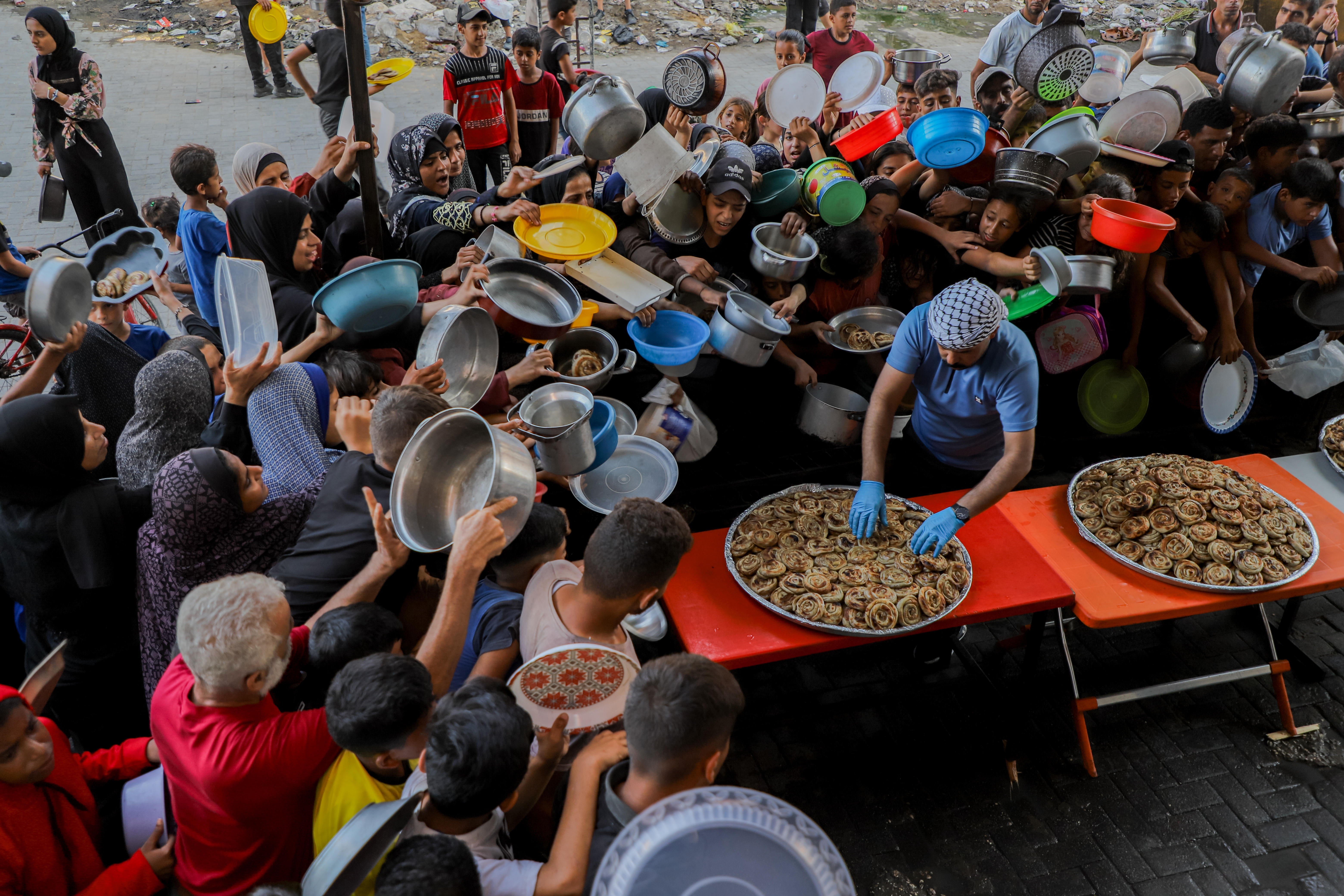 Eine große Gruppe von Menschen mit leeren Tellern, Schüsseln und anderen Gefäßen drängt sich um einen Helfer, der Essen aus einer großen Pfanne ausgibt.