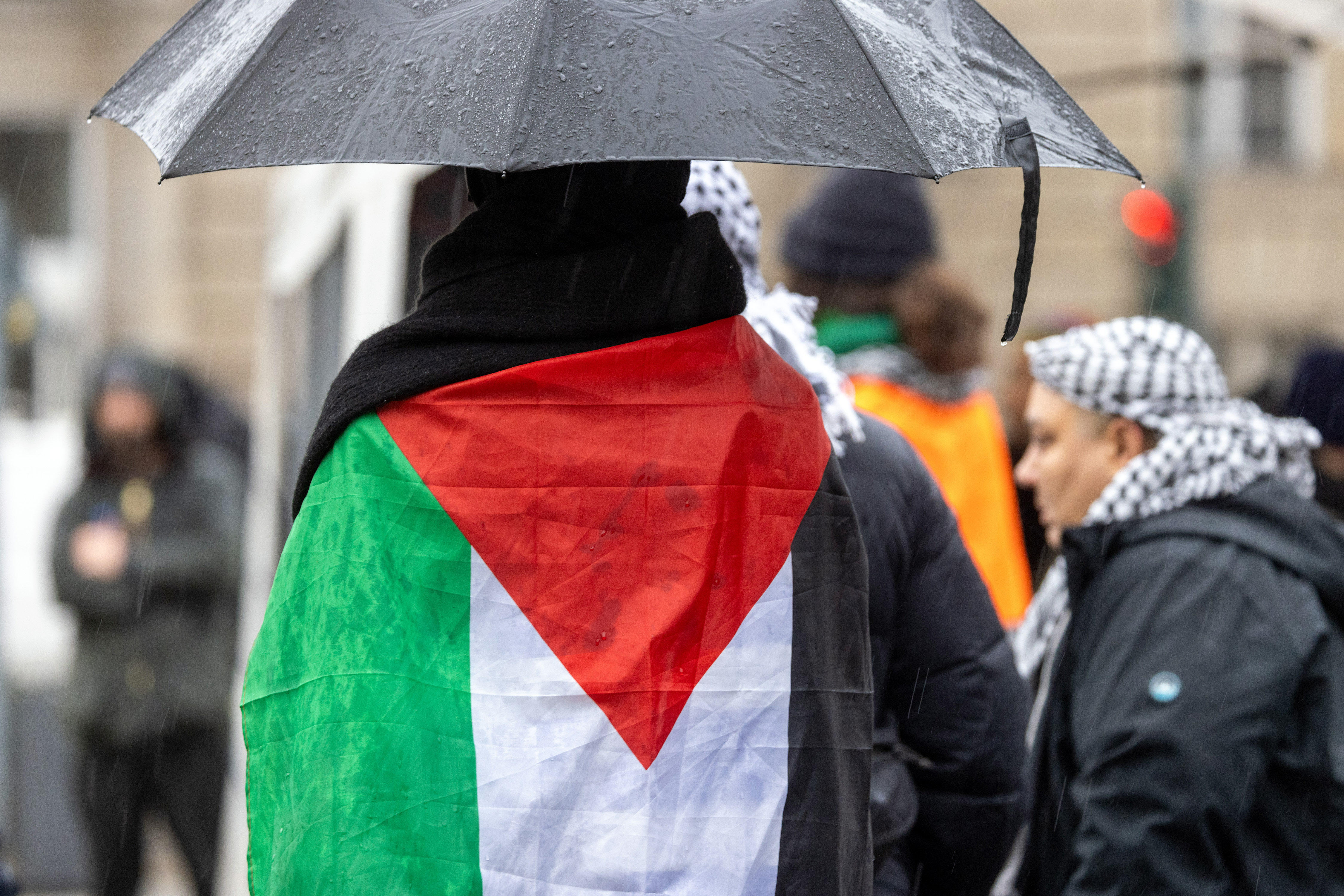 One person stands with their back to the camera, carrying an umbrella and with a Palestinian flag around their shoulders.