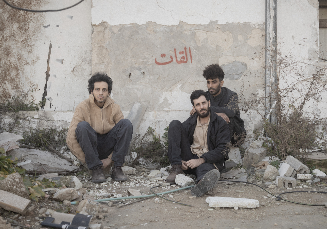 Three members of El Khat sit on the ground and on pieces of rubble in front of a white wall.