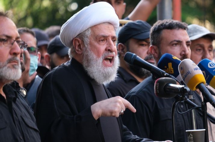 Naim Qassem wearing a white turban speaks into a microphone at a podium surrounded by men. His finger is pointing to the ground.