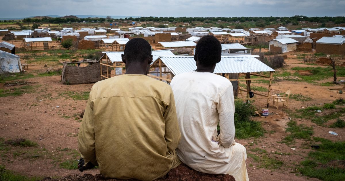 Two men sit facing away from the camera, looking over a field of makeshift shelters built from timber and corrogated iron.