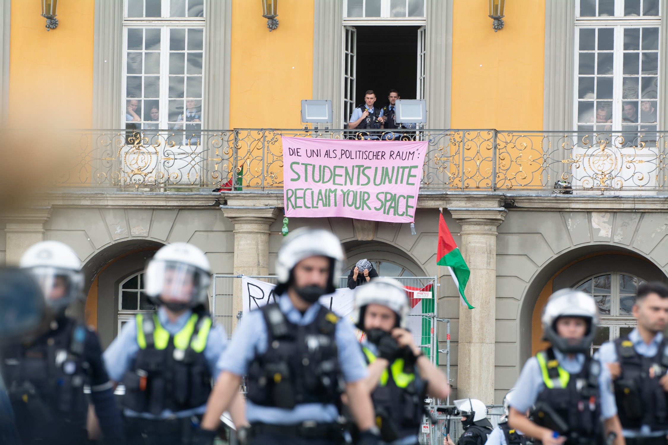 A group of police stand in front of a large University building wearing helmets. In the background, a Palestine flag and a banner hangs from the building that reads: "Students Unite. Reclaim Our Space."