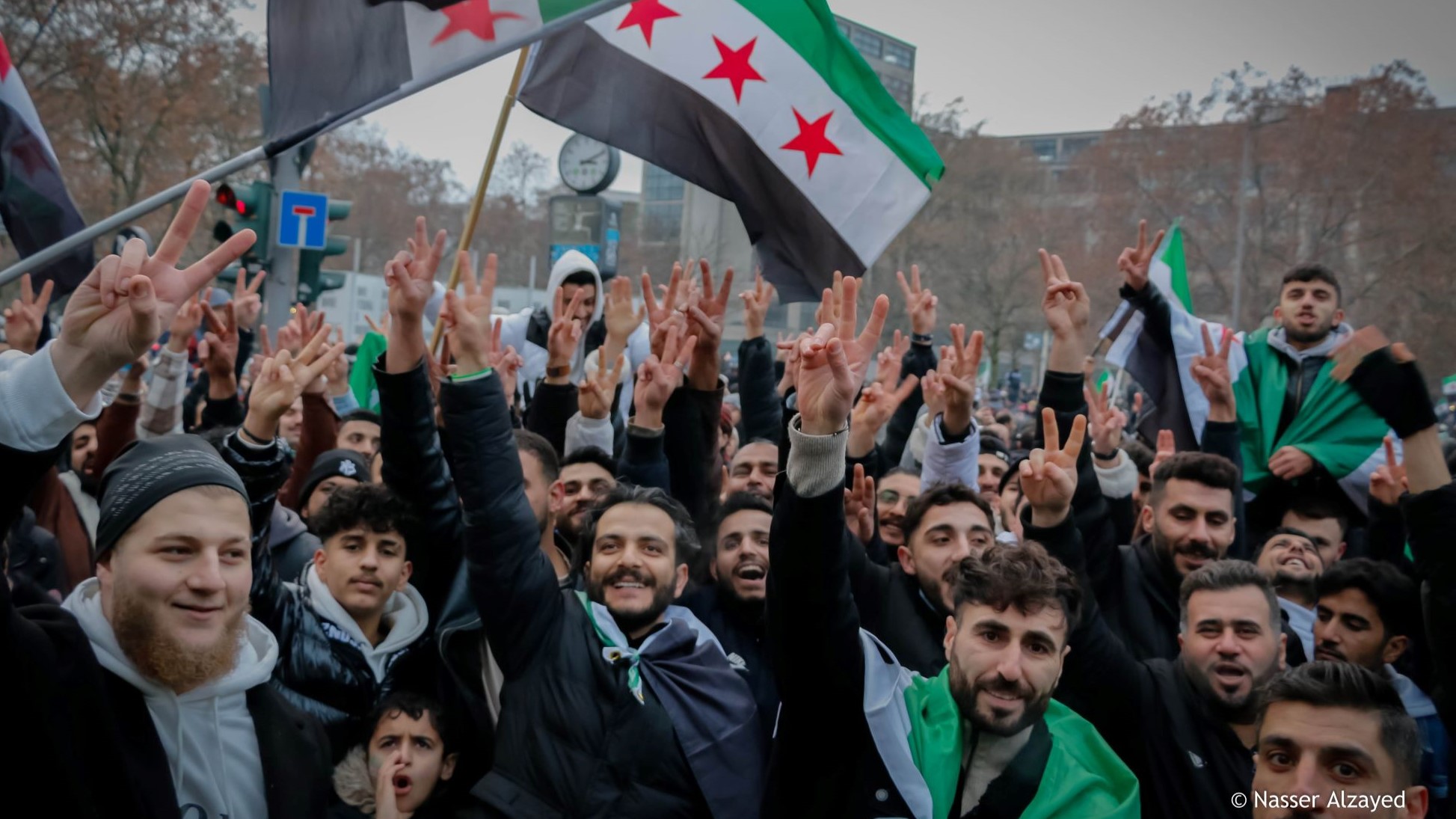 A crowd of people make peace signs to the camera. Syrian revolutionary flags wave in the background.