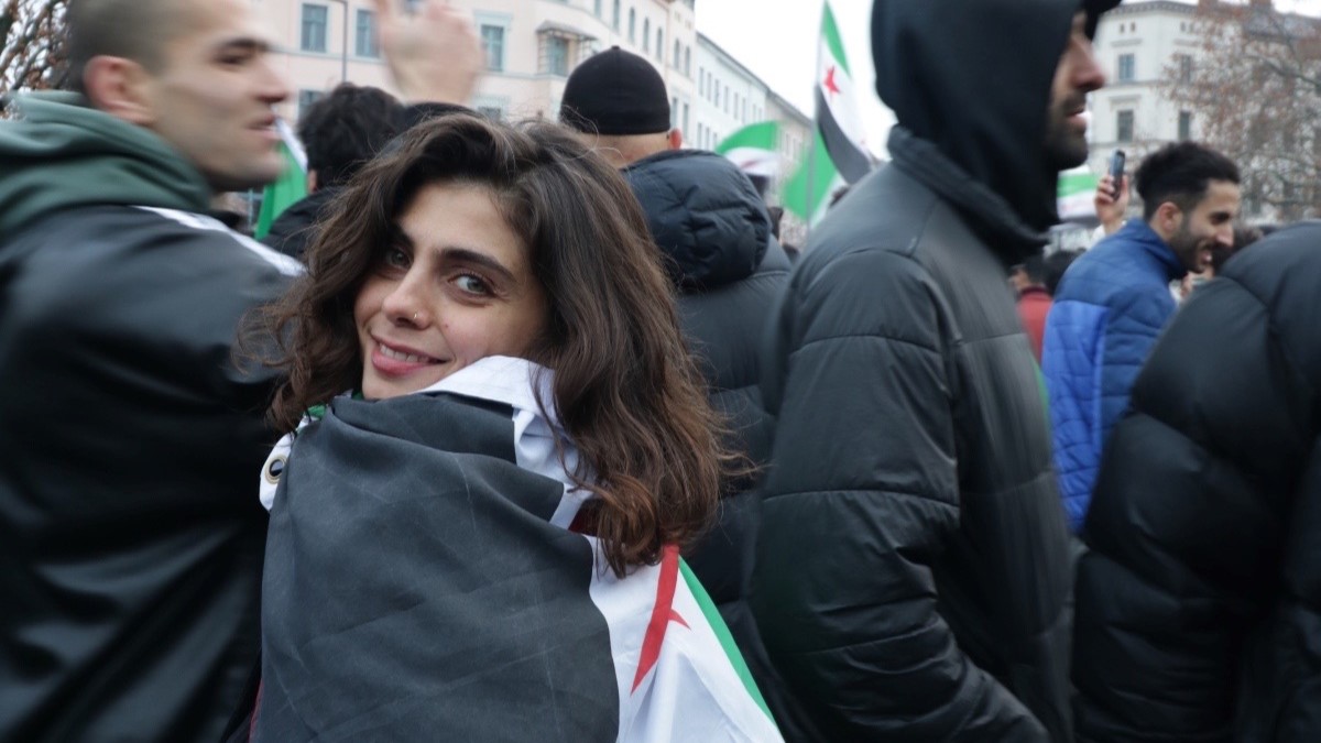 A woman wears a flag at a protest. She smiles at the camera.