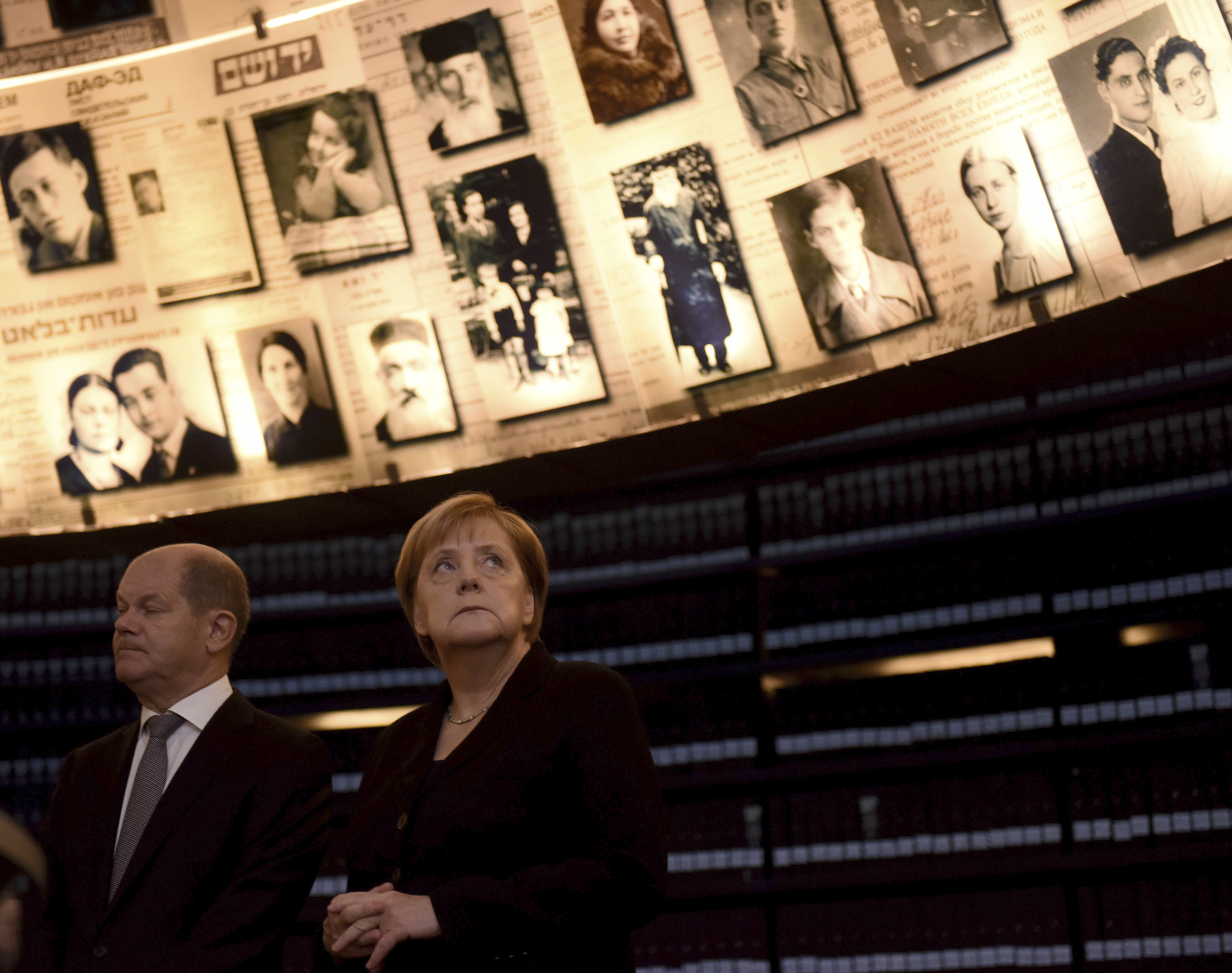 Merkel and Olaf Scholz stand next to eachother, on the wall behind them are a number of black and white photographs.