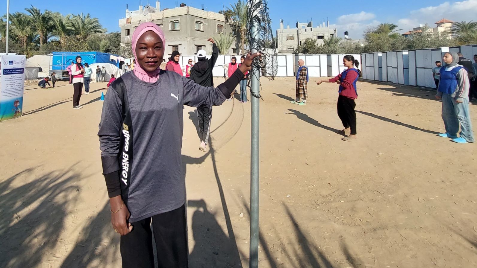 A woman in a pink headscarf and sportswear stands on a dirt court.