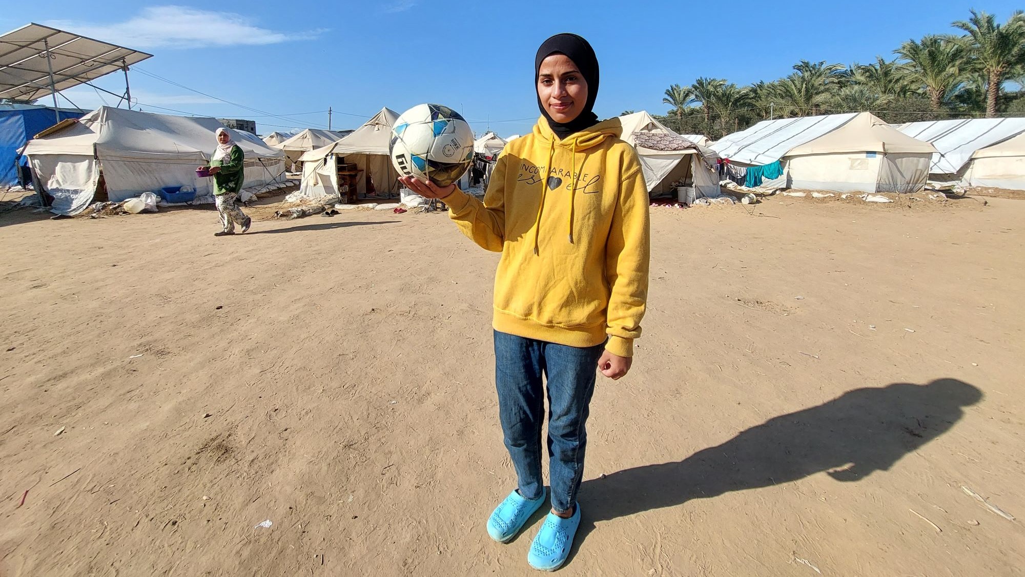 A woman in a headscarf and yellow hoodie holds a volleyball. In the background are tents.