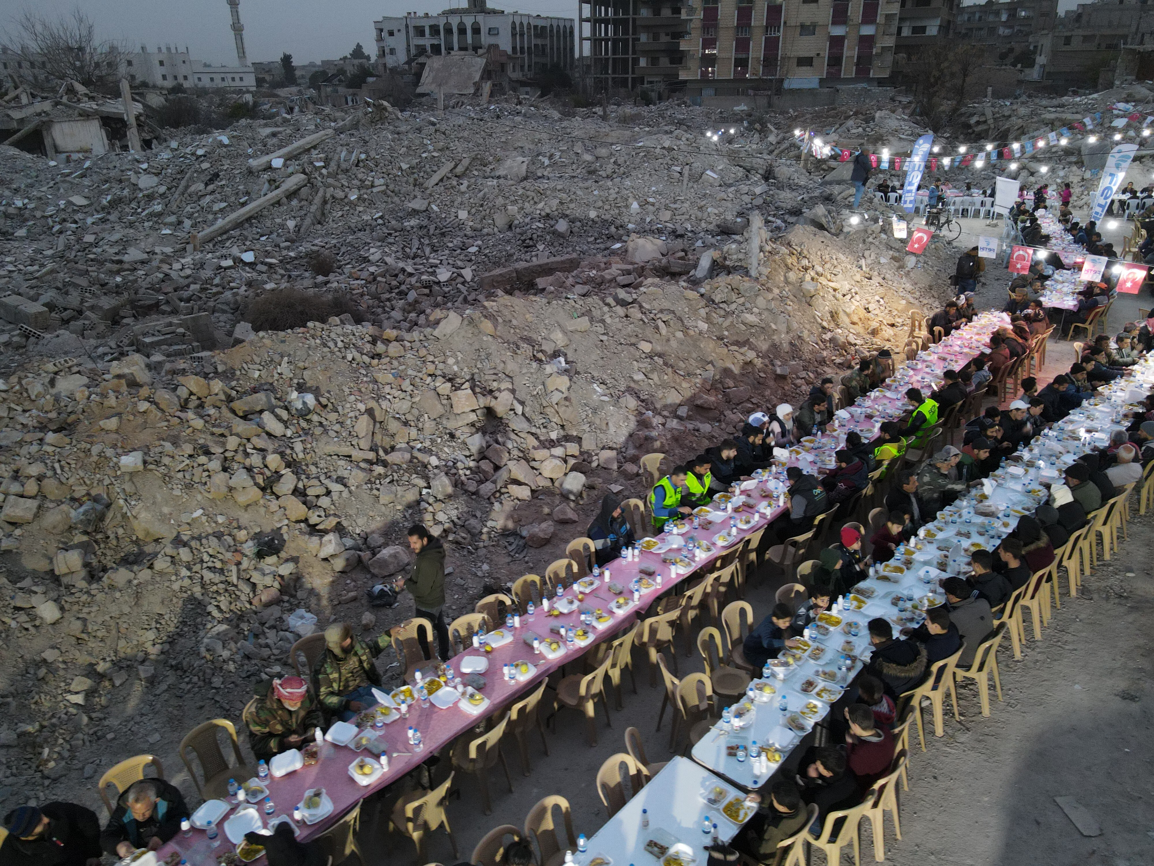 A long table with plastic chairs among ruins. Some people sit on chairs.