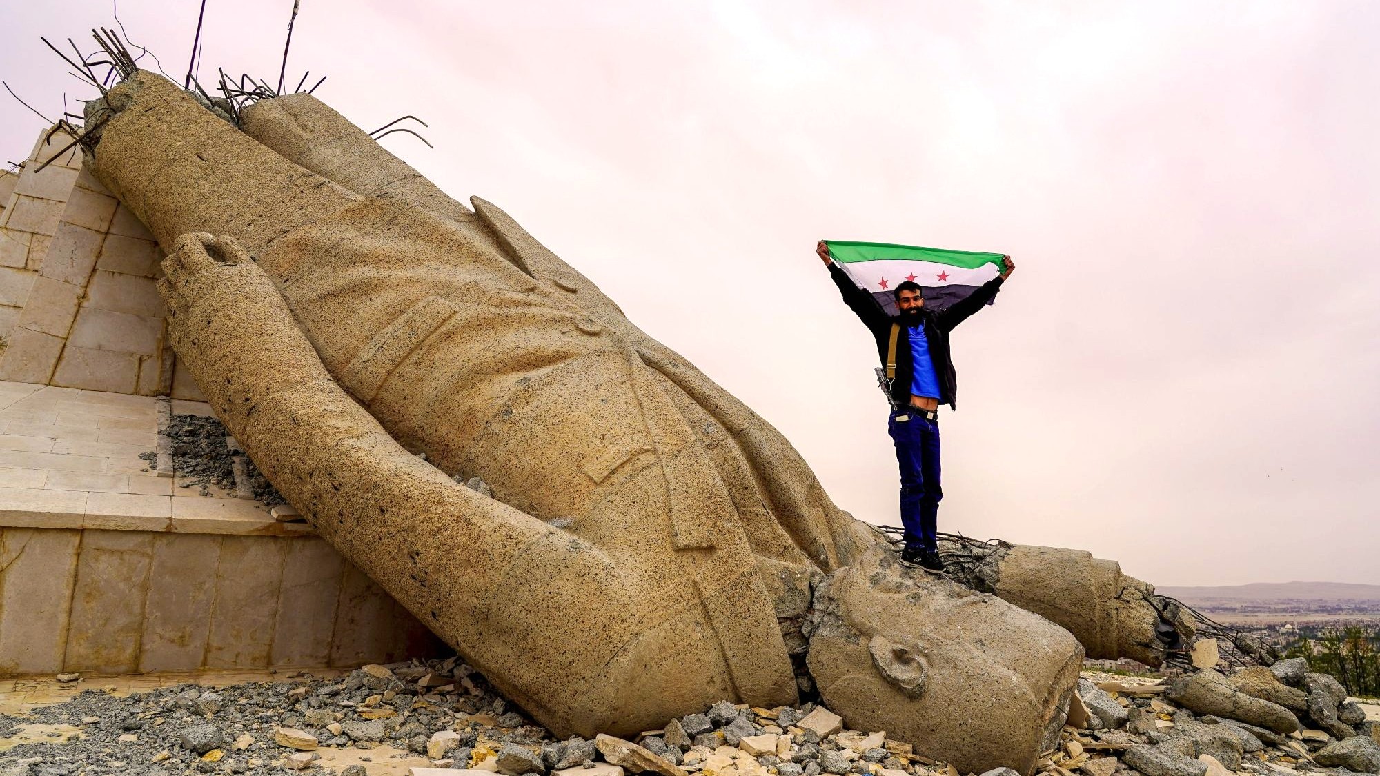 Deir Atiye in the Damascus countryside, January 2025 Picture Alliance / SIPA | L. Barakat A man with the flag of free Syria stands on the head of a toppled Assad statue.