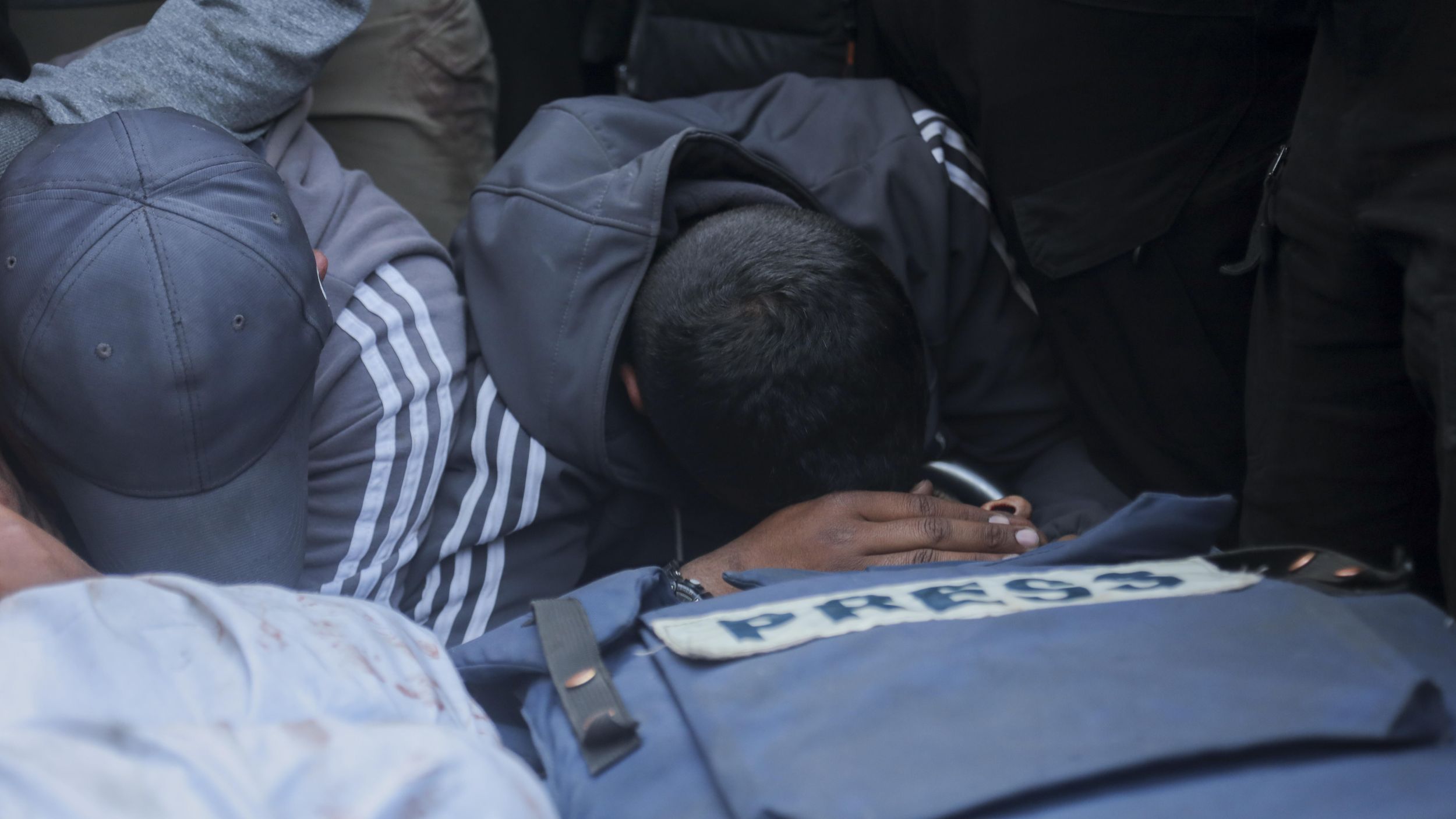 Family and friends mourn Hossam Shabat in Gaza, March 2025. (Photo: picture alliance / Middle East Images | A. Abosalm)a Two men kneel and bow their heads over a blue press vest.