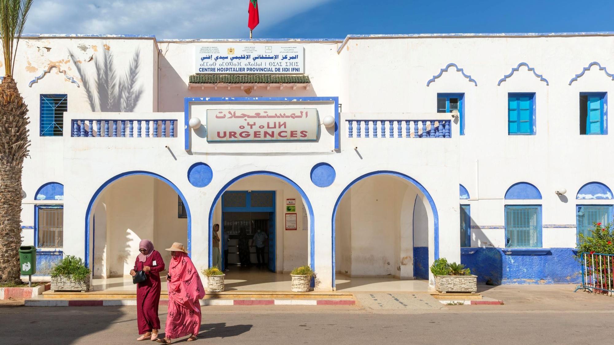 امرأتان تسيران أمام مبنى مستشفى يعود إلى الاستعمار الإسباني في سيدي إفني، المغرب، نوفمبر/تشرين الثاني 2022. (Photo: Picture Alliance / imageBROKER | Ian Murray) امرأتان بزي تقليدي تسيران خارج مبنى مستشفى.